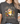 A woman wearing a dark gray graphic t-shirt with a cartoon sheep character and the text 'SORRY TO THE 99 I was dilly dallying'.