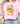 Person wearing a pink t-shirt with decorative text and graphics, sitting next to flowers.