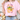 Person wearing a pink t-shirt with decorative text and graphics, sitting next to flowers.