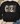 A black crewneck sweatshirt laid flat on a light wood surface, featuring the word 'COZY' in large letters filled with holiday patterns including snowflakes, plaid, polka dots, and snowmen. Beige sneakers and blue jeans are partially visible around it, with eucalyptus leaves in the corners.
