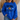 A woman wearing a royal blue crewneck sweatshirt with a black and brown football graphic and text on the front, paired with blue jeans in a cozy indoor setting.