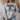 A woman wearing a light gray crewneck sweatshirt with a black and brown football graphic and text on the front, paired with light blue jeans in a cozy indoor setting.