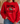 A person wearing a bright red crewneck sweatshirt with a black and white football graphic on the front, paired with blue jeans in a cozy indoor setting.