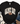 A black short-sleeve graphic T-shirt laid flat with a distressed white-and-black polka-dot arch reading “GAME DAY” and a brown football graphic in the center. The shirt is styled with blue jeans, a knit blanket, and a brown boot on a white wood background.