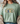A woman wearing a sage green t-shirt with a decorative 'Grace' graphic on the chest, styled with a beige knit cardigan and blue jeans.