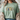 A woman wearing a sage green t-shirt with a decorative 'Grace' graphic on the chest, styled with a beige knit cardigan and blue jeans.
