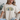 A woman wearing a beige graphic t-shirt with a floral 'Grace' design, sitting on a light-colored couch.