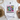 A white short-sleeve graphic T-shirt laid flat on a light surface with decorative props around it. The shirt features a colorful smiley face design with flowers, pink text reading 'Be happy it drives people crazy,' and black-and-white checkered borders.
