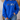 A person wearing a bright royal blue crewneck sweatshirt with a graphic of four cartoon sports figures on the chest, paired with blue jeans in a softly lit indoor setting.