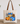 A beige canvas tote bag with brown shoulder straps, featuring a colorful illustration of patterned pumpkins and flowers on the front, displayed on a wooden surface against a white wall with greenery in the background.
