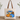A beige canvas tote bag with brown shoulder straps, featuring a colorful illustration of patterned pumpkins and flowers on the front, displayed on a wooden surface against a white wall with greenery in the background.