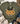An olive green graphic t-shirt with the text 'but GOD' in black and orange, decorated with pink flowers, styled in a flat-lay with jeans, a hat, boots, and a throw blanket.
