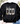A black crewneck sweatshirt laid flat on a light wood surface, featuring the text 'but GOD' across the chest, with 'GOD' filled in a camouflage pattern. The sweatshirt is styled with eucalyptus leaves, white flowers, a pair of white sneakers, and blue jeans.