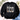 A black crewneck sweatshirt laid flat on a light wood surface, featuring the text 'but GOD' across the chest, with 'GOD' filled in a camouflage pattern. The sweatshirt is styled with eucalyptus leaves, white flowers, a pair of white sneakers, and blue jeans.