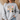 A woman wearing a light gray crewneck sweatshirt with a graphic print of nine cute, patterned chickens in various colors, paired with blue jeans in a cozy indoor setting.