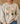 A person wearing a beige crewneck sweatshirt with a colorful graphic of nine cartoon chickens in various patterns, paired with blue jeans in a cozy indoor setting.