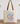 A beige canvas tote bag with brown shoulder straps, featuring a colorful graphic of a stethoscope and flowers on a striped background, with the text 'Created with Purpose' printed below. The bag is displayed on a wooden surface against a white wall with decorative plants in the background.
