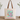 A beige canvas tote bag with brown shoulder straps, featuring a colorful graphic of a stethoscope and flowers on a striped background, with the text 'Created with Purpose' printed below. The bag is displayed on a wooden surface against a white wall with decorative plants in the background.