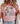 A woman wearing a light pink graphic T-shirt with a floral and cross design that reads 'Daughter of the King' in black text. She is also wearing light blue jeans and a cream knit cardigan, with blonde hair and a leafy plant visible in the background.