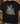A black short-sleeve graphic t-shirt featuring a detailed print of three deer in a forest, styled in a flat-lay on a rustic wood background with hunting accessories.
