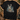 A black short-sleeve graphic t-shirt featuring a detailed print of three deer in a forest, styled in a flat-lay on a rustic wood background with hunting accessories.