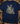 A navy blue short-sleeve t-shirt with a detailed graphic of three deer in a forest, laid flat on a rustic wooden surface surrounded by hunting-themed props like a camouflage cap, binoculars, and a boot.