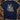 A navy blue short-sleeve t-shirt with a detailed graphic of three deer in a forest, laid flat on a rustic wooden surface surrounded by hunting-themed props like a camouflage cap, binoculars, and a boot.