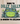 A cream canvas tote bag with brown shoulder straps sits on a wooden surface against a white wall. The bag features a printed graphic of a green and blue frog with the text 'BORN TO DILLY DALLY & KNOWN TO LOLLYGAG'.
