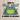 A cream canvas tote bag with brown shoulder straps sits on a wooden surface against a white wall. The bag features a printed graphic of a green and blue frog with the text 'BORN TO DILLY DALLY & KNOWN TO LOLLYGAG'.