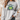 A woman wearing a light gray crewneck sweatshirt with a graphic of a green frog and the text 'BORN TO DILLY DALLY & KNOWN TO LOLLYGAG'.