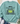 A mint green crewneck sweatshirt laid flat on a white wood background, styled with blue jeans, white sneakers, a straw hat, and eucalyptus leaves. The sweatshirt features a graphic of a green toad with a yellow belly and the text 'BORN TO DILLY DAILY & KNOWN TO LOLLYGAG'.