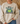 A person wearing a beige crewneck sweatshirt with a graphic of a green frog and the text 'BORN TO DILLY DALLY & KNOWN TO LOLLYGAG'.