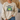 A person wearing a beige crewneck sweatshirt with a graphic of a green frog and the text 'BORN TO DILLY DALLY & KNOWN TO LOLLYGAG'.