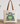 A cream canvas tote bag with brown shoulder straps sits on a wooden surface against a white wall. The bag features a printed graphic of a green and blue frog with the text 'BORN TO DILLY DALLY & KNOWN TO LOLLYGAG'.