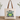 A cream canvas tote bag with brown shoulder straps sits on a wooden surface against a white wall. The bag features a printed graphic of a green and blue frog with the text 'BORN TO DILLY DALLY & KNOWN TO LOLLYGAG'.