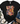 A black short-sleeve graphic T-shirt laid flat on a white wood background, styled with a cream throw blanket, greenery, blue jeans, and brown lace-up boots. The shirt features a colorful patchwork-style 'LOVE LIKE DOLLY' text design with butterflies and leopard print details.