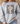 A woman wearing a light gray crewneck sweatshirt with a graphic print that reads 'FAITH FAITH FAITH' in distressed black letters, overlaid with a cream and black polka dot bow design, paired with light blue jeans.