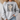 A woman wearing a light gray crewneck sweatshirt with a graphic print that reads 'FAITH FAITH FAITH' in distressed black letters, overlaid with a cream and black polka dot bow design, paired with light blue jeans.