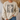 A person wearing a cream-colored crewneck sweatshirt with a graphic print that reads 'FAITH FAITH FAITH' in distressed black letters, overlaid with a gift box design featuring a beige polka-dot ribbon. The person is also wearing blue jeans, standing in a cozy indoor setting with a couch and dried palm leaves in the background.
