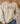 A person wearing a cream-colored crewneck sweatshirt with a graphic of four cute marshmallot characters standing in a row. The background shows a cozy indoor setting with a beige couch and dried pampas grass.