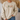 A person wearing a cream-colored crewneck sweatshirt with a graphic of four cute marshmallot characters standing in a row. The background shows a cozy indoor setting with a beige couch and dried pampas grass.
