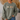 A person wearing an olive green crewneck sweatshirt with a graphic of four small cartoon characters on the chest, paired with blue jeans in a cozy indoor setting.