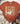 A rust-orange graphic t-shirt featuring a white goose wearing a plaid bow and heart-shaped sunglasses while holding an iced coffee, styled in a flat-lay with jeans, a knit blanket, and boots on a white wood background.