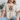 A woman wearing a beige graphic t-shirt with a raccoon design and the text 'feral girl autumn' in orange and black, paired with ripped blue jeans, sitting on a couch.