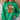 A person wearing a bright green crewneck sweatshirt with a large gingerbread cookie graphic wearing a leopard-print Santa hat. The background shows a neutral indoor setting with a woven straw chair.