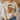 A person wearing a cream-colored crewneck sweatshirt with a large graphic of a winking gingerbread cookie wearing a leopard-print Santa hat and glasses, paired with blue jeans.