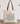 A beige canvas tote bag with brown leather handles, featuring a large star graphic on the front with the phrase 'God is good all the time' in script and patterned star details, set on a wooden surface against a white wall with eucalyptus branches.