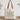 A beige canvas tote bag with brown leather handles, featuring a large star graphic on the front with the phrase 'God is good all the time' in script and patterned star details, set on a wooden surface against a white wall with eucalyptus branches.