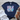 Navy blue t-shirt with 'God is Good all the time' text on a wooden surface with white jeans and brown sandals.