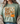 A woman wearing a sage green graphic t-shirt with a fall-themed design that reads 'God is Good ALL THE TIME' alongside a pumpkin and flowers, styled with a beige knit cardigan.